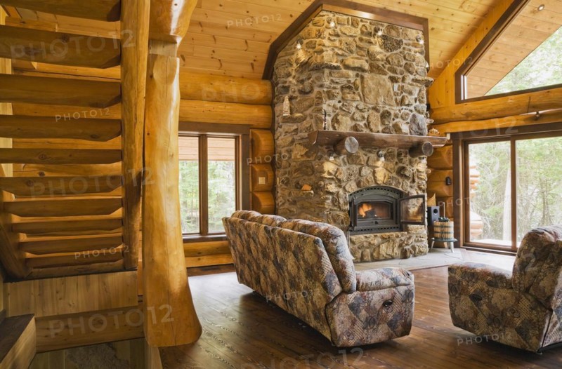 Wooden log stairs and upholstered sofas with lit fieldstone and porous rock fireplace in living room inside handcrafted red cedar log cabin home, Quebec, Canada, North America