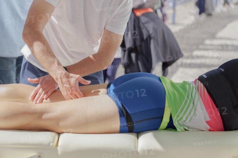 A first responder is administering first aid to a marathon runner who has collapsed on the ground. The rescuer is pressing with both hands on the runners back, possibly performing a massage or assessing the injury. The runner, dressed in athletic gear, lies prone on a white mat, suggesting a quick response to an incident during the event