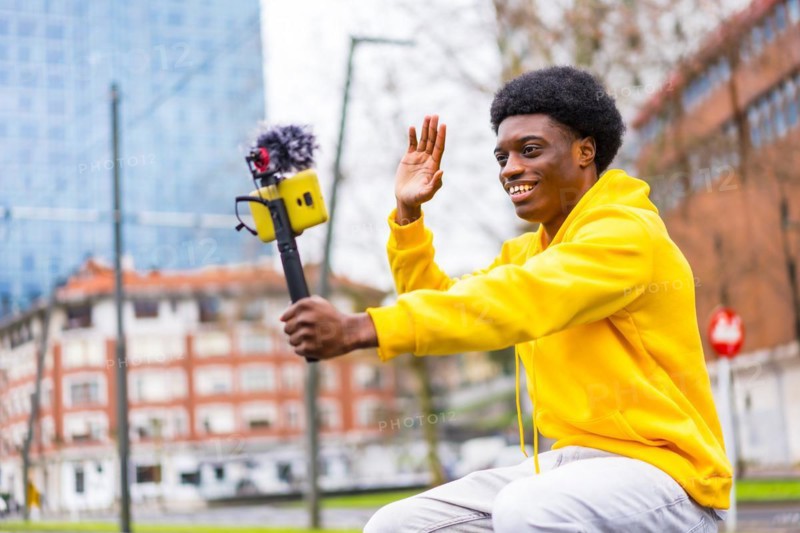 Side view photo of a young african stylish vlogger recording video and waving in the city