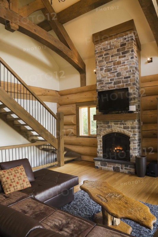Brown leather L-shaped sofa and wooden coffee table in front of lit nuanced tan and brown natural stone propane gas fireplace next to wooden stairs with wrought iron handrail leading to upstairs floor in living room with hardwood floor inside luxurious contemporary Scandinavian style log cabin home, Quebec, Canada, North America
