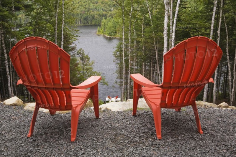 Two bright red plastic Adirondack chairs overlooking a calm lake through Betula, Birch trees with green leaves from gravel surface pation in backyard of a secluded log cabin home in late summer, Quebec, Canada, North America