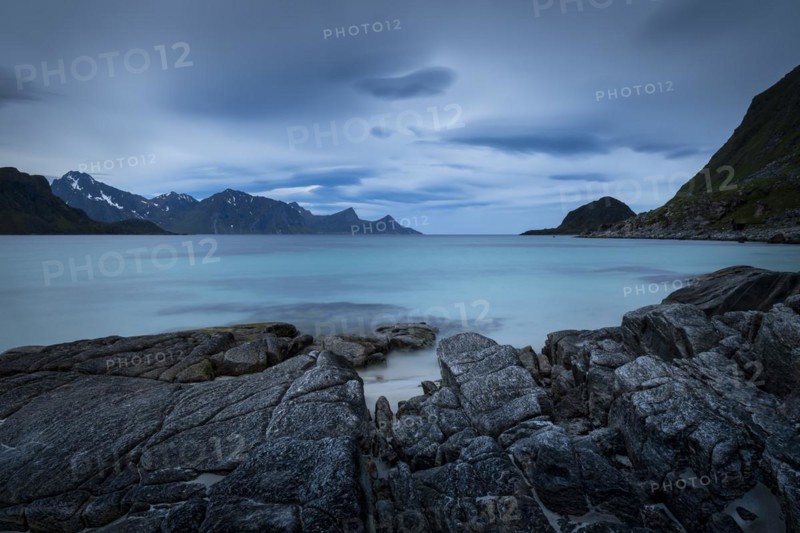 Seascape on the beach at Haukland. View of the mountains of Myrland on Flakstadoya. Dark sky, blurred clouds. Rocks in the foreground. Early summer. Long exposure. Haukland Beach, Haukland, Vestvagoya, Lofoten, Norway, Europe