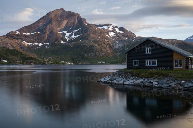 Landscape on the Lofoten Islands. A wooden house in Kakern on Kakersundet. The mountain Narvtinden on Moskenesoya in the background. The mountain and the house are reflected in the sea. At night at the time of the midnight sun in good weather, a few clouds in the sky. Early summer. Kakern, Flakstadoya, Lofoten, Norway, Europe