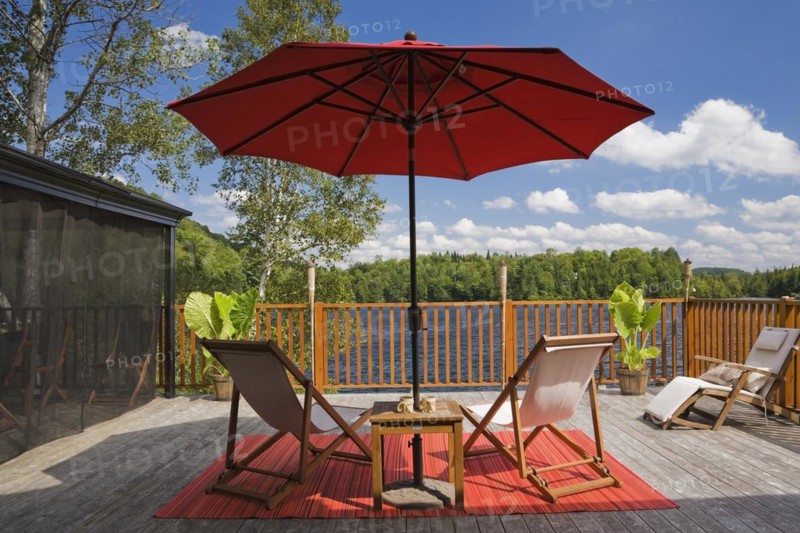 Black fishnet gazebo with red canvas cloth parasol and wooden folding canvas chairs on red mat on grey wood plank deck with railing overlooking lake with green forest in backyard in summer, Quebec, Canada, North America