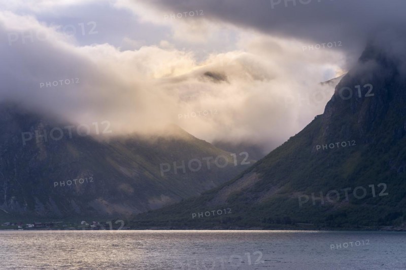 View over the Flakstadpollen to mountains in clouds with incoming sunlight. The small village of Flakstad in front. At night during the midnight sun. Early summer. Flakstadoya, Lofoten, Norway, Europe