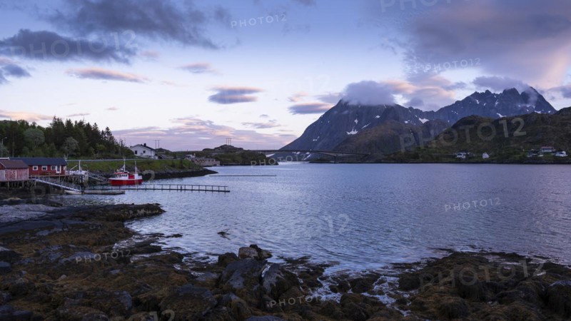 Landscape on the Lofoten Islands near Ramberg. On the left a few houses by the sea and a boat. Further back the Flakstad bridge (Flakstadbruene) and mountains on Moskenesoya. At night during the midnight sun. Good weather, some lenticular clouds in the sky. Early summer. Flakstadoya, Lofoten, Norway, Europe