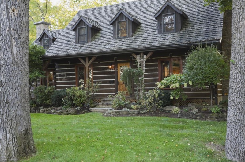 White chinked log cabin home facade with grey weathered cedar shingles roof, three dormer windows and landscaped front yard with mixed perennial shrubs and plants in borders in autumn, Quebec, Canada, North America