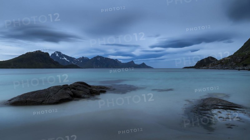 Seascape on the beach at Haukland. View of the mountains of Myrland on Flakstadoya. Dark sky, blurred clouds. Rocks in the foreground. Early summer. Long exposure. Haukland Beach, Haukland, Vestvagoya, Lofoten, Norway, Europe