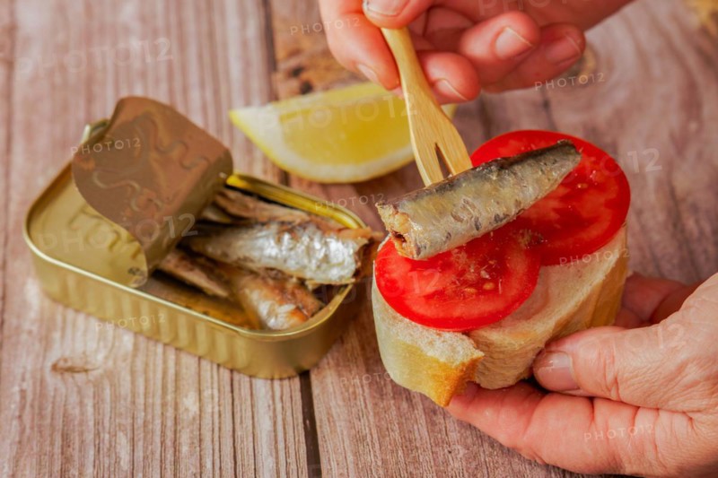 A woman puts a sardine on a slice of bread with tomato, in the background you can see the can of sardines and a lemon on a wooden table