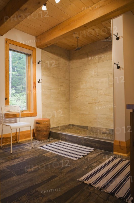 Illuminated beige ceramic shower stall in main bathroom with tan and dark grey ceramic tile floor and blue and white striped rugs on ground floor inside luxurious contemporary timber and milled log cabin home, Quebec, Canada, North America