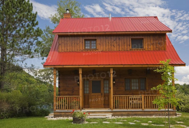 Small two story brown stained log cabin home with veranda and red sheet metal roof and landscaped front yard with deciduous tree and flagstone footpath in summer, Quebec, Canada, North America