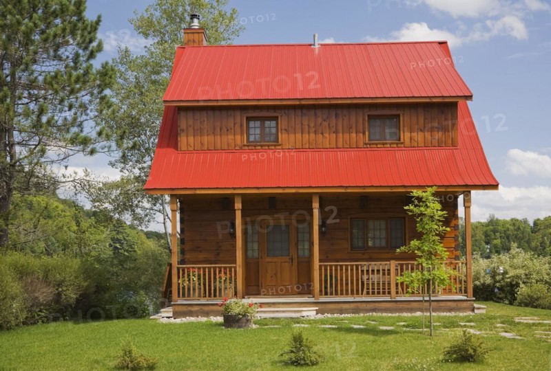 Small two story brown stained log cabin home with veranda and red sheet metal roof and landscaped front yard with deciduous tree and flagstone footpath in summer, Quebec, Canada, North America