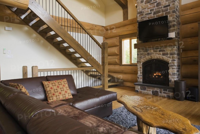 Brown leather L-shaped sofa and wooden coffee table in front of lit nuanced tan and brown natural stone propane gas fireplace next to wooden stairs with wrought iron handrail leading to upstairs floor in living room with hardwood floor inside luxurious contemporary Scandinavian style log cabin home, Quebec, Canada, North America