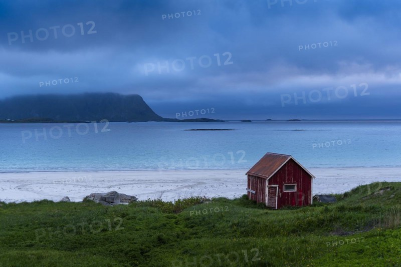 A red wooden hut on the sandy beach of Ramberg (Rambergstranda), sea and mountains. At night at the time of the midnight sun. Clouds in the sky. Early summer. Ramberg, Flakstadoya, Lofoten, Norway, Europe