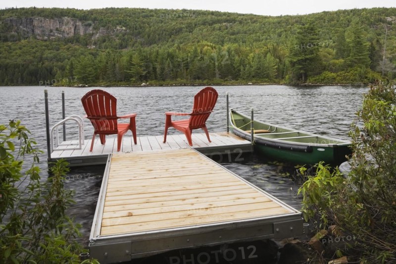 Two bright red plastic Adirondack chairs on floating wooden dock and green rowboat on calm lake with forest of green leaved coniferous and deciduous trees in late summer, Quebec, Canada, North America