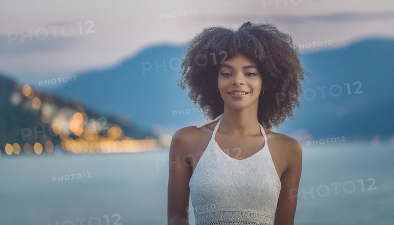 Smiling woman wearing a summer dress by the waterfront at sunset, radiating serenity, blurry moody landscaped background with bokeh effect, AI generated