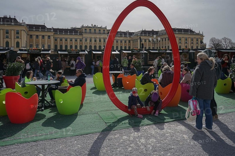 Lively Easter market with children and adults sitting on colourful apple chairs, with a large red symbolised Easter egg in the background Schoenbrunn Palace Vienna