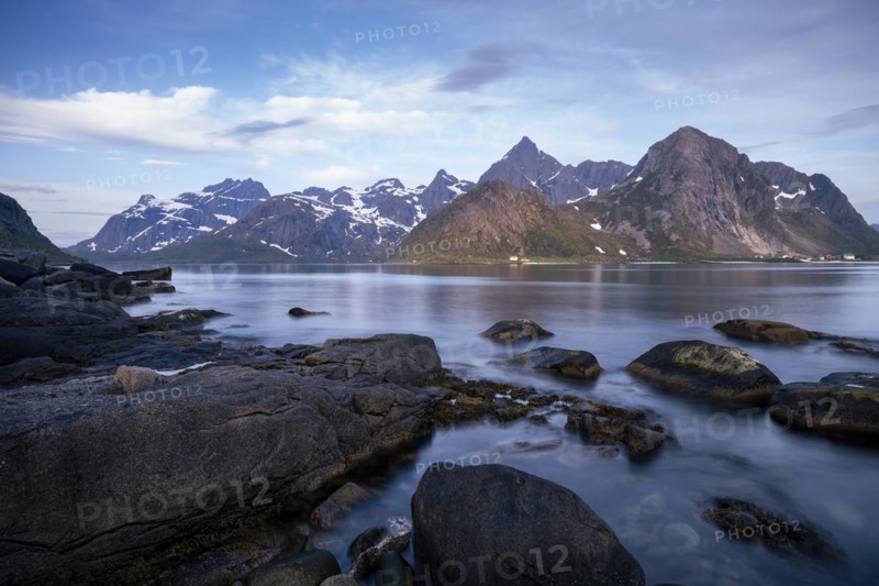 Landscape with sea and mountains on the Lofoten Islands, view over the fjord Flakstadpollen to the small village Flakstad and the mountains Flakstadtinden, Stortinden and other mountains. Rocks in the foreground. At night at the time of the midnight sun in good weather, some clouds in the sky. Long exposure. Early summer. Flakstadoya, Lofoten, Norway, Europe