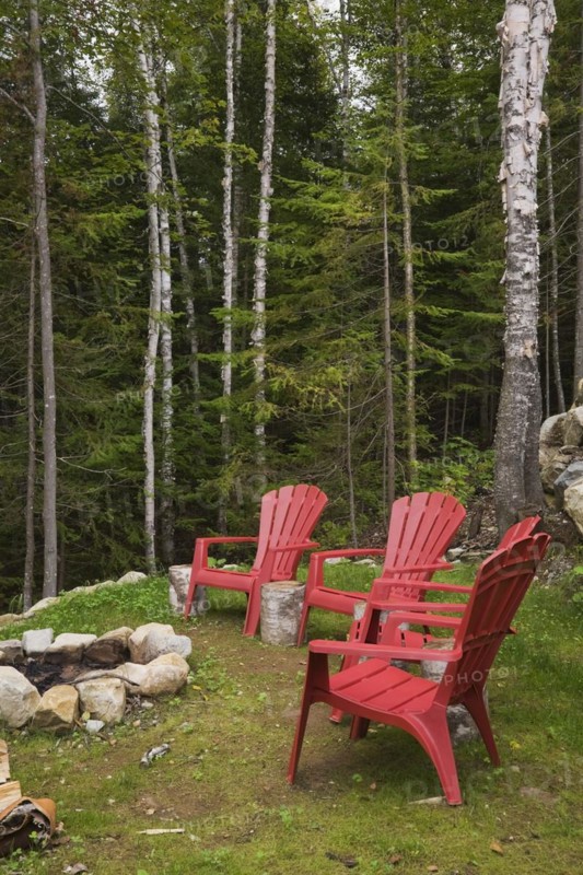 Four bright red plastic Adirondack chairs around rock edged firepit dug into the ground in backyard surrounded by a forest of evergreen and deciduous trees in late summer, Quebec, Canada, North America