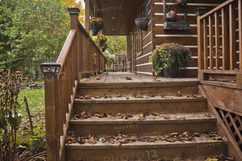 Old and worn brown stained wooden stairs and long veranda with pine wood rocking chair and fallen tree leaves on facade of rustic white chinked log cabin home in autumn, Quebec, Canada, North America