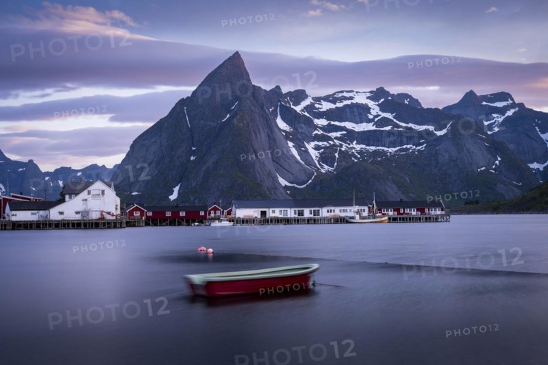 Landscape on the Lofoten Islands. The village of Hamnoy. A small boat in the foreground, blurred by long exposure. Houses and Mount Olstinden in the background. At night at the time of the midnight sun in good weather, some clouds in the sky. Early summer. Hamnoy, Moskenesoya, Lofoten, Norway, Europe
