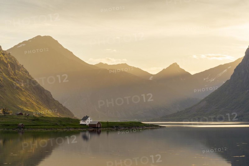 Landscape with sea and mountains on the Lofoten Islands. The fjord Flakstadpollen and some houses belonging to the village of Flakstad. The landscape is reflected in the sea. At night at the time of the midnight sun in good weather, the sun shines in from the side. Flakstadoya, Lofoten, Norway, Europe