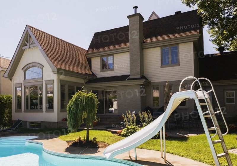 Rear view of a grey brick and cream cladding with grey trim contemporary cottage style home and landscaped backyard with in-ground swimming pool and slide in summer, Quebec, Canada, North America