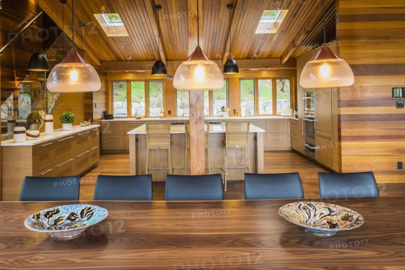 Illuminated industrial style copper with frosted glass pendant lighting fixtures over wooden dining table with black leather chairs plus kitchen with bamboo wood island and cabinets in background inside luxurious stained cedar and timber wood home with panoramic windows, Quebec, Canada, North America