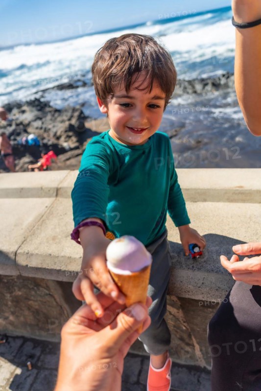 A young boy is holding an ice cream cone and giving it to another person. The scene is set on a beach, with the ocean in the background. Scene is lighthearted and playful