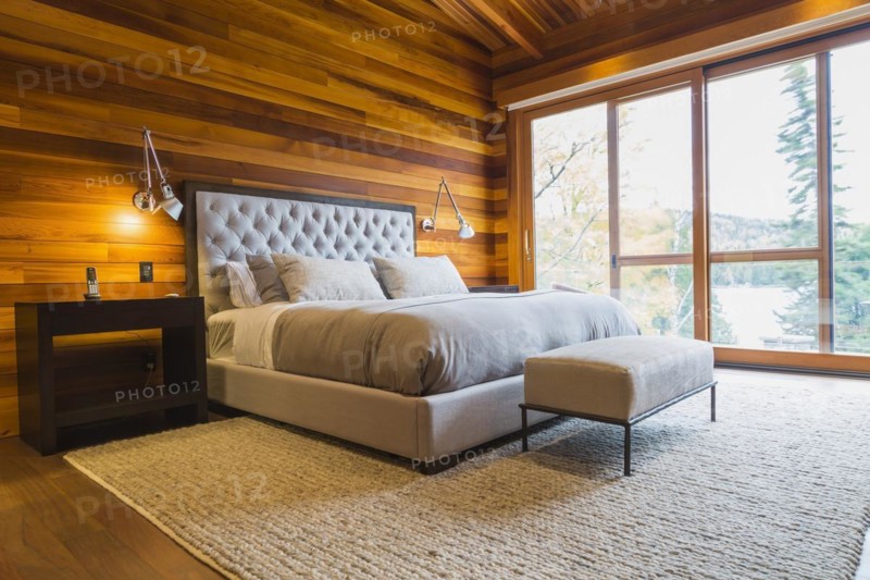 King size bed with grey upholstered panel headboard, ottoman and wooden end tables in master bedroom with grey nuanced rug and Ipe wood floor inside luxurious stained cedar and timber wood home with panoramic windows, Quebec, Canada, North America