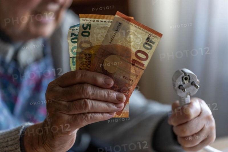 Senior citizen holding money and a power cable with plug in her hand at home, symbolising energy costs and poverty, Cologne, North Rhine-Westphalia, Germany, Europe