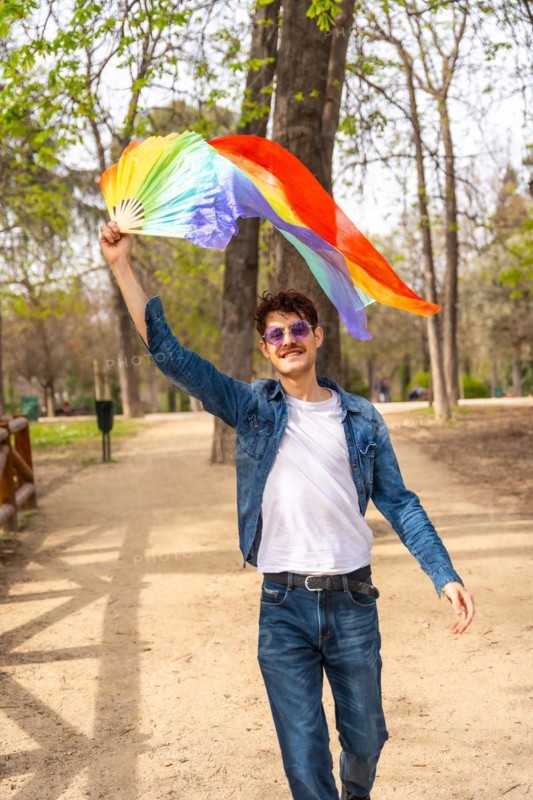 Vertical photo of a gay man raising and waving a rainbow colored hand fan in a park