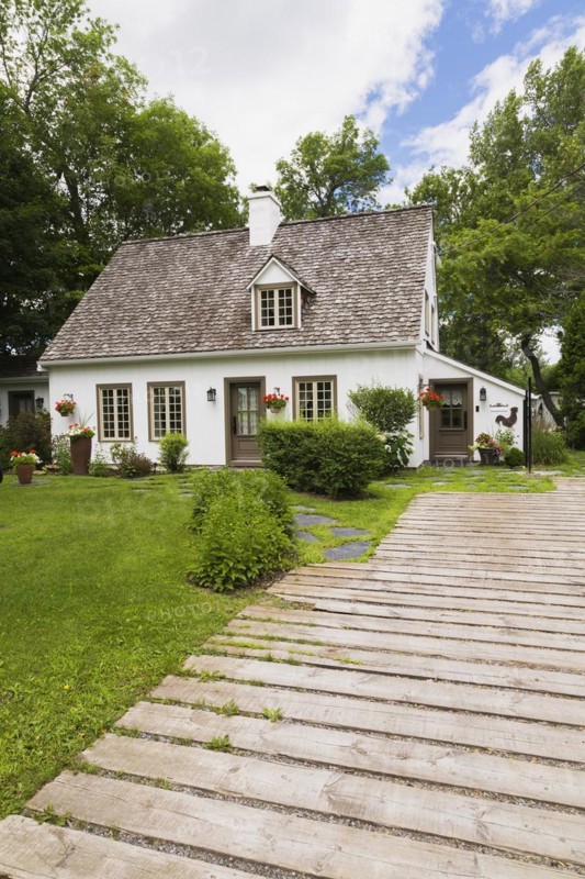 Old circa 1886 white with beige and brown trim Canadiana cottage style home facade and wood plank driveway in summer, Quebec, Canada, North America