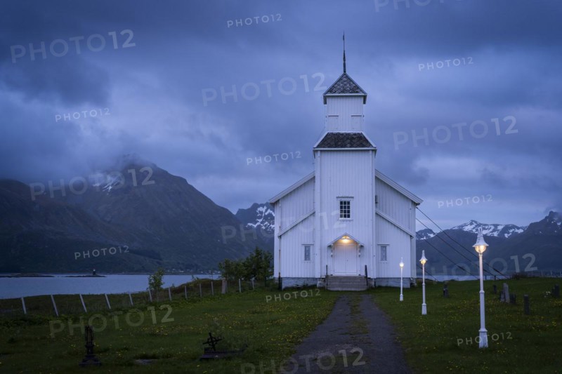 The white church of Gimsoy (Gimsoysand church) by the sea. A path leads to the church. The church and lanterns are illuminated. At night at the time of the midnight sun. Cloudy sky. Early summer. Gimsoya, Lofoten, Norway, Europe