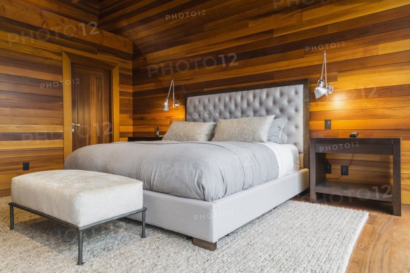King size bed with grey upholstered panel headboard, ottoman and wooden end tables in master bedroom with grey nuanced rug and Ipe wood floor inside luxurious stained cedar and timber wood home, Quebec, Canada, North America