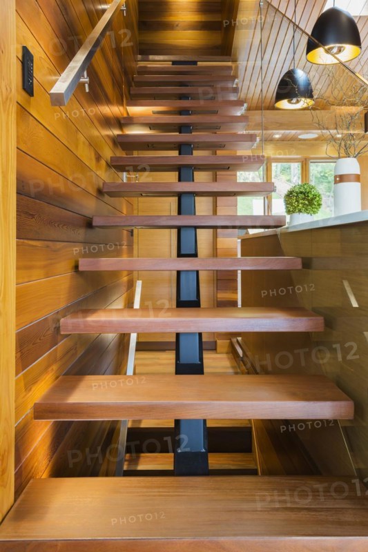 Looking up illuminated wooden staircase with opened steps and clear glass railing leading to upstairs floor inside luxurious stained cedar and timber wood home, Quebec, Canada, North America
