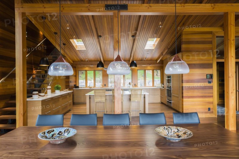 Industrial style copper with frosted glass pendant lighting fixtures over wooden dining table with black leather chairs plus kitchen with bamboo wood island and cabinets in background inside luxurious stained cedar and timber wood home with panoramic windows, Quebec, Canada, North America