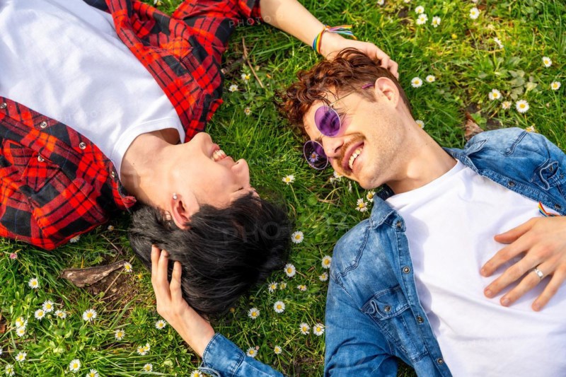 Top view photo of a multi-ethnic gay couple lying on the grass of a park together looking each other and smiling