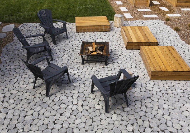 Grey paving stone patio with black Adirondack sitting chairs, slightly raised flat rectangular red cedar wooden bench platforms around a metal firepit in residential backyard in summer, Quebec, Canada, North America