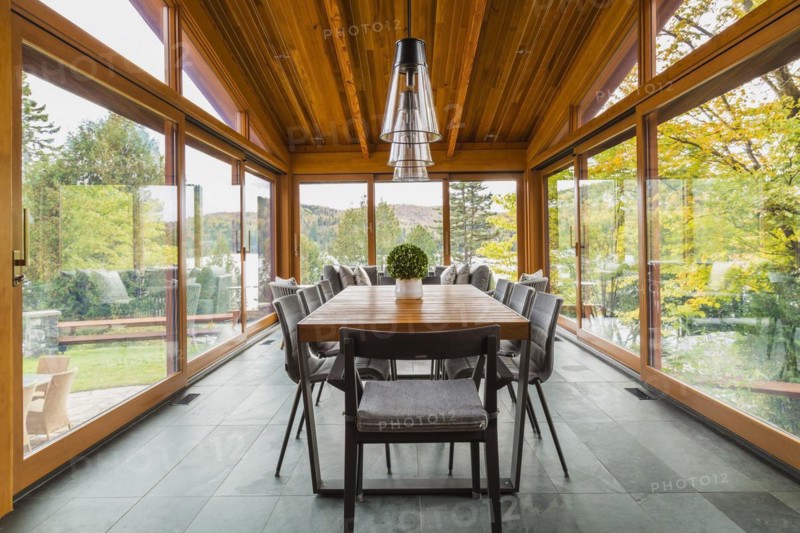 Four season gazebo and industrial style black with clear glass pendant lighting fixtures over wood and black metal dining table with grey upholstered chairs and heated slate floor inside luxurious stained cedar and timber wood home with panoramic windows, Quebec, Canada, North America