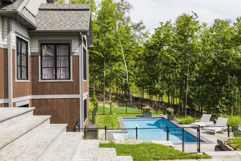 In-ground swimming pool and rear view of contemporary natural stone and brown stained wood and cedar shingles clad luxurious bungalow in summer, Quebec, Canada, North America
