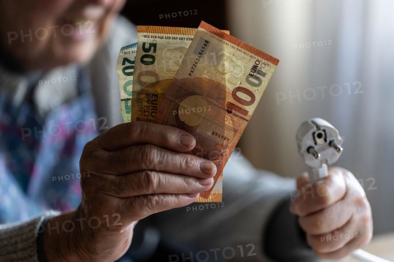 Senior citizen holding money and a power cable with plug in her hand at home, symbolising energy costs and poverty, Cologne, North Rhine-Westphalia, Germany, Europe
