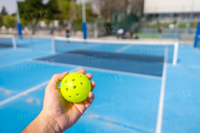 Unrecognizable person holding a plastic yellow pickleball ball with blue outdoor court on the background