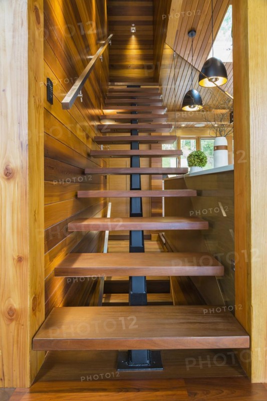 Looking up illuminated wooden staircase with opened steps and clear glass railing leading to upstairs floor inside luxurious stained cedar and timber wood home, Quebec, Canada, North America