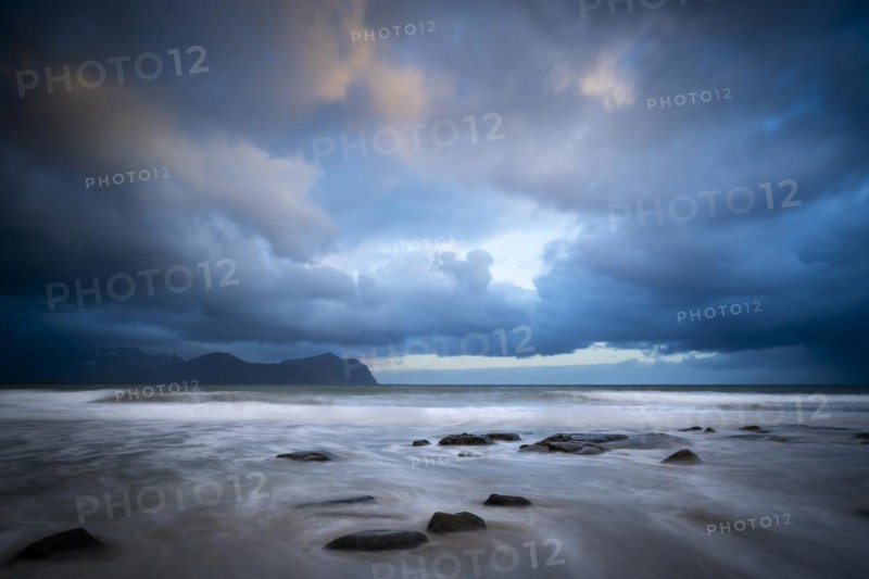 On the beach at Vikten. The sea washes around some rocks. Mountains in the background. At night at the time of the midnight sun. Cloudy, dramatic sky. Early summer. Long exposure. Vikten, Flakstadoya, Lofoten, Norway, Europe