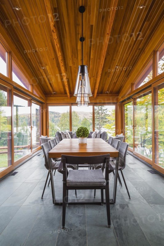 Four season gazebo and illuminated industrial style black with clear glass pendant lighting fixtures over wood and black metal dining table with grey upholstered chairs and heated slate floor inside luxurious stained cedar and timber wood home with panoramic windows, Quebec, Canada, North America