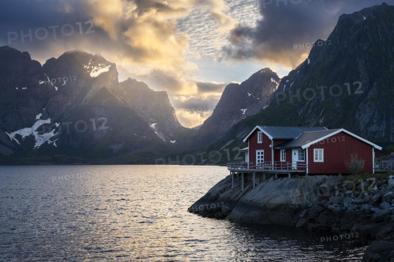 A typical red wooden house on wooden stilts (rorbuer) in the small village of Toppoya (Toppoy), overlooking the sea and the mountains. At night at the time of the midnight sun. Clouds and blue sky, the sun shines on a mountain peak. Toppoya, Moskenesoya, Lofoten, Norway, Europe