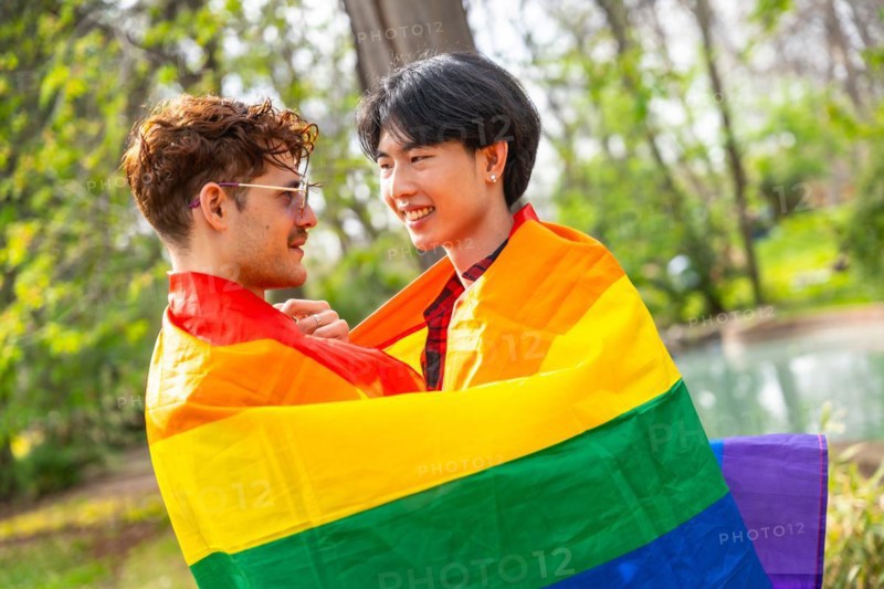 Multi-ethnic gay couple embraced in a park wrapped in rainbow flag