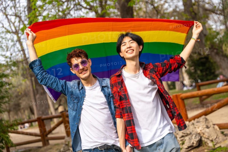 Portrait of a multi-ethnic gay couple raising a rainbow lgbt flag in a park