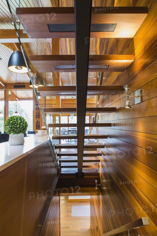 Underside view of illuminated wooden staircase with opened steps and clear glass railing inside luxurious stained cedar and timber wood home, Quebec, Canada, North America